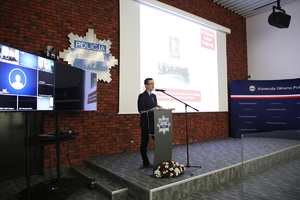 Mężczyzna przemawia do uczestników seminarium stojąc przy mównicy, w tle na ścianie gwiazda policyjna, po lewej stronie zestaw do wideokonferencji